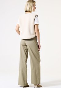 Top sin mangas beige y peludo sobre una camisa blanca, combinado con pantalones de pierna ancha verde oliva y zapatos de tacón marrón claro. Diseño minimalista.