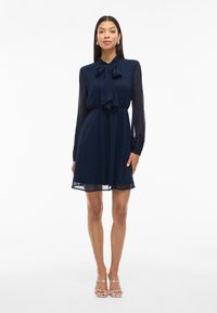 Γυναίκα που στέκεται με το πρόσωπο στραμμένο μπροστά, φορώντας ένα ναυτικό μπλε φόρεμα με μακριά μανίκια και μια δεμένη κορδέλα στον λαιμό, συνδυασμένο με ασημένιες ανοιχτές γόβες.