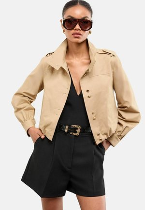 Beige cropped jacket with gold buttons, paired with a black deep V-neck top and tailored black shorts. Black textured belt accents the outfit.