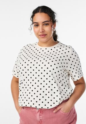 Žena s tmavými kučeravými vlasmi nosí bielu košeľu s krátkym rukávom s čiernymi bodkami a ružové nohavice, ruky v kapsách, na bielom pozadí.