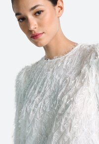 Jauna sieviete ar dabīgu grimu, valkājot baltu, fakturētu, spalvu apģērbu, skatās nedaudz uz leju vienkārša gaiša fona priekšā.