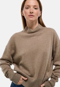 Femme avec des cheveux bruns lisses portant un pull beige ample à col haut et un jean bleu, posant avec les mains dans les poches.