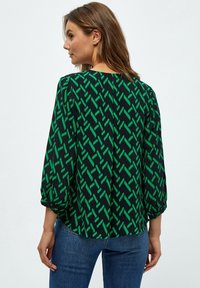 Čierna blúzka s zeleným geometrickým vzorom, vo voľnom strihu, s tromi štvrtinovými rukávmi a zaobleným lemom, skombinovaná s modrými džínsami.