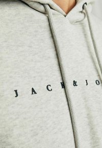 Sudadera ligera gris claro hecha de una tela suave que cuenta con una capucha ajustable y "JACK&JONES" impreso en letras negras en negrita en el pecho.