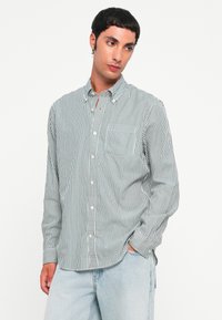 Chemise à rayures vert foncé et blanc en coton, avec un col, des manches longues et une poche poitrine. Portée avec un jean bleu clair.