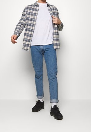 Camisa de cuadros azul y blanca con botones, superpuesta sobre una camiseta blanca, combinada con vaqueros azules remangados y zapatillas negras.