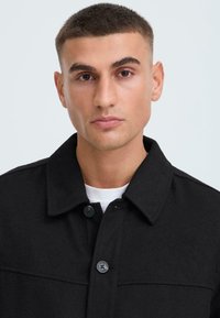 Zwarte wolmix jas met een knoopkraag, voorzien van twee knopen aan de voorkant en een gestructureerde stof. Draagt over een wit shirt.