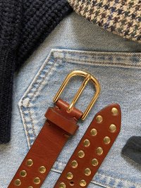 Brunt läderbälte med ett guldfärgat dubbelt spänne; detaljerat med mässingsnitar på en rem, mot en bakgrund av denimtyg och stickad textur.