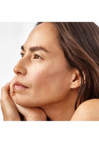 Profil av en kvinna med medelbrunt hår, ljus hy, definierade kindben och subtila fräknar nära näsan, som vilar hakan på sina händer.