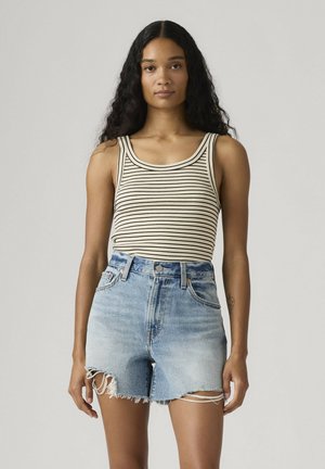 Mujer con cabello largo y rizado, vestida con una camiseta sin mangas de rayas beige y negra y unos shorts de mezclilla azul claro de cintura alta con bordes deshilachados.