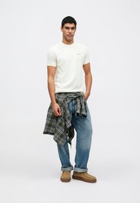 Κρεμ κοντομάνικο μπλουζάκι, μπλε τζιν σε χαλαρή εφαρμογή, καρό φανέλα δεμένη γύρω από τη μέση και ταμπά παπούτσια τύπου slip-on. Απλή, καθημερινή εμφάνιση.