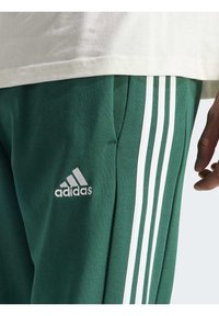 Gröna träningsbyxor med tre vita ränder längs sidan och en broderad Adidas-logga. Tillverkade av texturerat tyg med fickor.