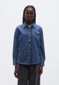 Tummansininen farkkupaita, jossa on terävä kaulus, napillinen suljenta, yksi rintatasku ja taskussa brändilaatta. Pitkät hihat.