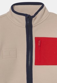 Fleecejack met een crèmekleurig lichaam, marineblauwe rits en kraag, en een rode zak met een vierkant ontwerp op de linkerborst.