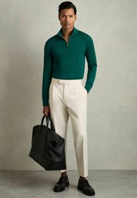 Un pull zippé vert, un pantalon crème ajusté, des chaussures noires et un sac fourre-tout en cuir noir. Texture lisse, design ajusté et forme structurée.