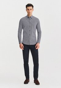 Mężczyzna stoi zrelaksowany, z rozłożonymi ramionami, ubrany w wzorzystą, długą koszulę z guzikami, ciemne spodnie i brązowe buty na jednolitym tle.