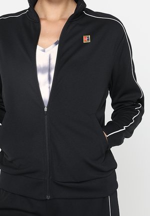 Person trägt eine schwarze Zip-up-Jacke mit weißen Paspeln an den Ärmeln und einem kleinen mehrfarbigen quadratischen Logo auf der Brust.