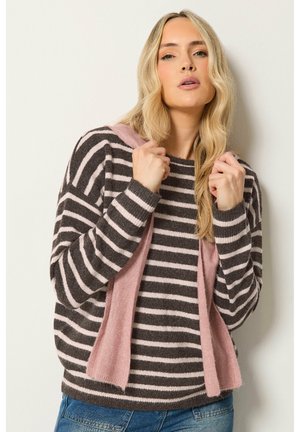 Frau mit langen blonden Haaren, die einen braun-pinken gestreiften Pullover trägt und einen pinken Schal über die Schultern drapiert hält.