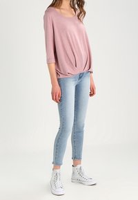 Hellrosa, locker sitzendes Hemd mit rundem Halsausschnitt und geknoteter Vorderseite, kombiniert mit hellblauen, schmal geschnittenen Jeans und weißen High-Top-Sneakern.