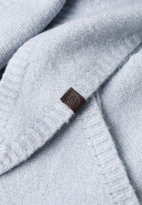 Material din lână texturată de culoare bleu deschis, cu un design pliat; prezintă o etichetă mică din piele maro cu un logo. Apariție moale și călduroasă.