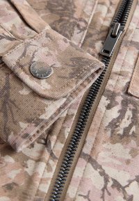 Gros plan d'un tissu camouflage beige avec une fermeture éclair noire et un bouton-pression métallique sur le rabat d'une poche de vêtement.