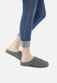 Pantofole grigie lavorate a maglia con un motivo a trecce testurizzato e design aperto sul retro, abbinate a jeans blu arrotolati, che mostrano un look informale.