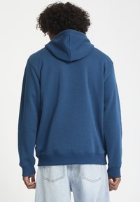 Blå hættetrøje lavet af blødt stof med en kengurulomme, ribbede manchetter og kant, samt en hætte med snor. Båret med lyse denimjeans.