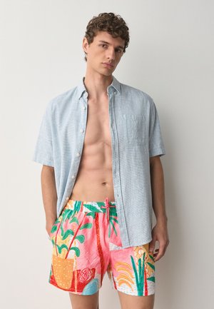 Jeune homme portant une chemise bleu clair à manches courtes ouverte sur un short imprimé tropical coloré, debout devant un fond clair uni.