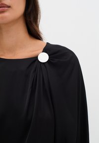 Top in tessuto nero drappeggiato con una fibbia circolare bianca testurizzata sulla spalla, creando una silhouette liscia ed elegante.