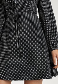 Schwarzes Kleid mit einem Sternenmuster in Weiß. Der Stoff ist leicht und hat einen figurschmeichelnden Schnitt mit einem verstellbaren Kordelzug in der Taille. Lange Ärmel.
