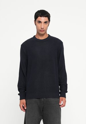 Jeune homme portant un pull en laine bleu marine foncé et un jean noir délavé, debout devant un fond blanc uni.