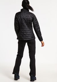 Schwarze gesteppte Jacke mit hohem Kragen und taillierter Form, kombiniert mit schwarzen Hosen und Wanderschuhen, von hinten gezeigt.