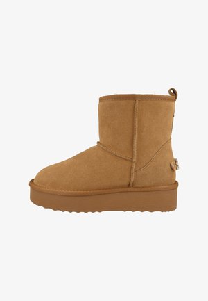 Stivaletti alla caviglia in camoscio color cognac con punta arrotondata, fodera in pelle di pecora, altezza alla caviglia e dettaglio decorativo con logo in metallo sul lato. Suola piatta in gomma.