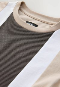 T-shirt beige et blanc avec un panneau côtelé texturé marron foncé. Il présente un col rond et l'étiquette de la marque est visible à l'intérieur.