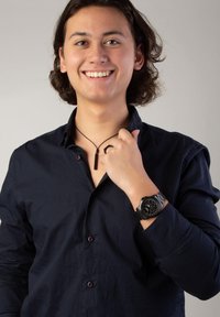 Schwarze Uhr mit rundem Ziffernblatt, Metallarmband; schwarzes Anhänger am Band; trägt ein dunkles Hemd mit Knöpfen; neutraler Hintergrund.