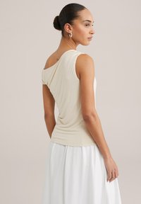 Bežajna, asimetrična bluza s kratkim rukavima s drapiranim dizajnom. Mekana, glatka tekstura; nosi se uz bijelu, lepršavu suknju. Elegantan i prilagođen silueta.