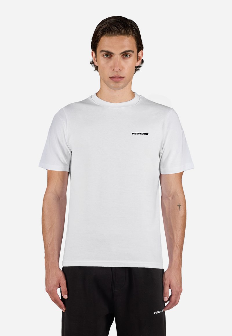 Giovane uomo in piedi rivolto in avanti, indossa una t-shirt Pegador bianca, pantaloni neri e ha un piccolo tatuaggio a croce sull'avambraccio sinistro.