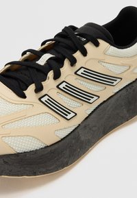 Baskets en mesh beiges avec lacets et accents noirs, dotées d'une semelle noire épaisse et de trois bandes noires sur le côté pour un détail supplémentaire.