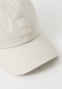 Texturált anyagból készült bézs sapka, varrott napellenzővel. Egy négyzet alakú folt található rajta, amelyen világos színű "BOSS" logó látható.