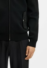 Zwarte gebreide zip-up jas met geribbelde manchetten en zoom. Bevat een gestructureerde zwarte, gewatteerde zak met een drukknoopdetail.