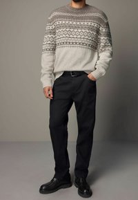Sweater in crème met een gemêleerde grijze schouderpartij. Gepaard met zwarte broek en stevige zwarte schoenen. Effen achtergrond met neutrale tinten.