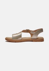Metallisk guld sandal med öppen tå och justerbart vristband, med en platt, texturerad brun sula och subtilt utskurna mönster.