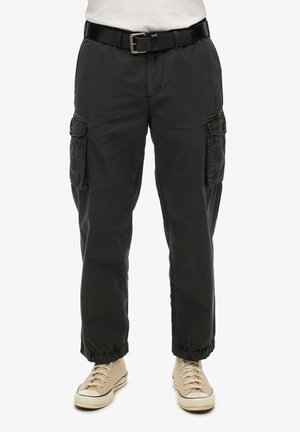 Pantaloni cargo neri con vestibilità rilassata, dotati di tasche laterali e orli a coste, abbinati a una cintura nera e sneaker beige.