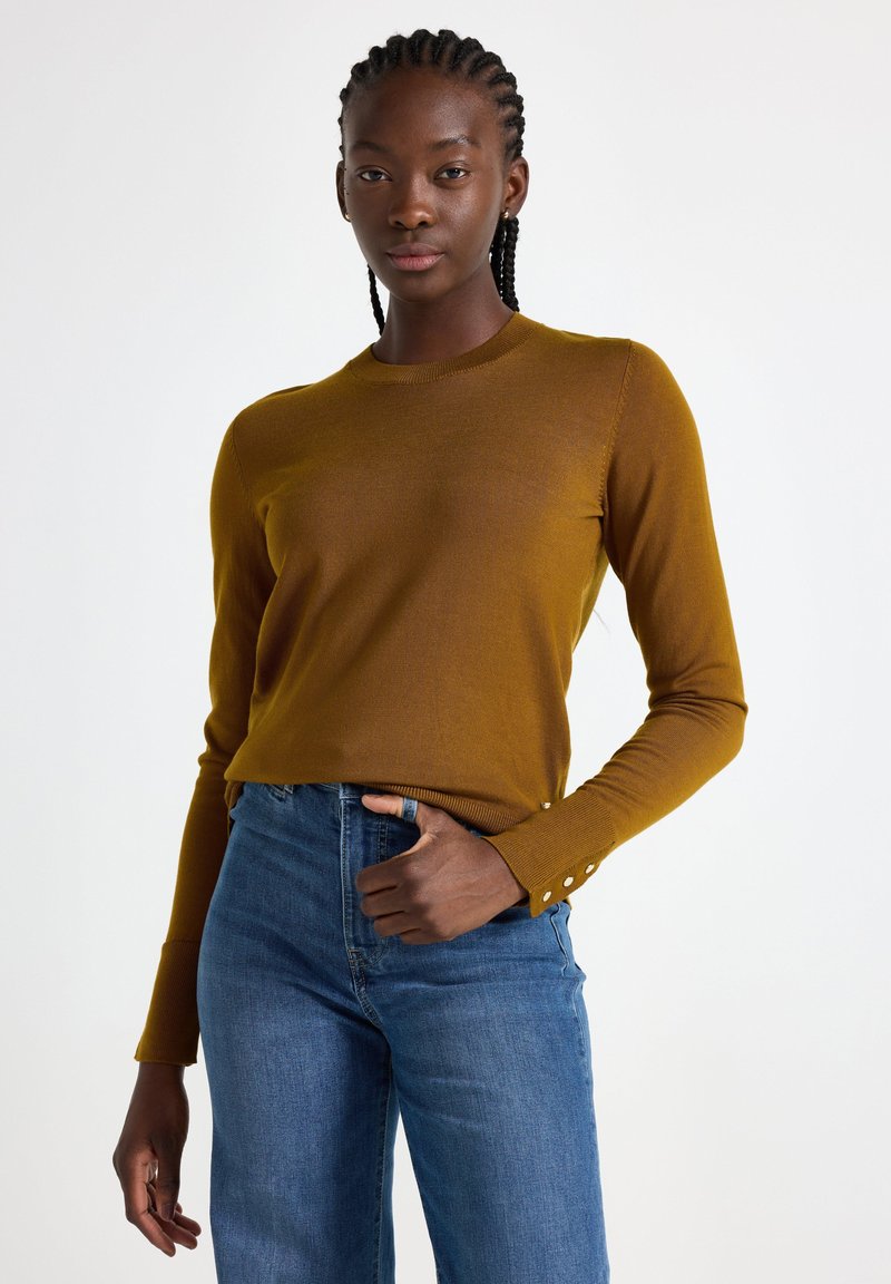 Senapsgul långärmad tröja med rund halsringning och knappdetaljer vid ärmsluten, tillsammans med blå denimjeans.