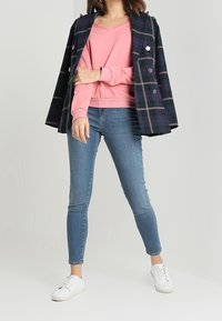 Donna con un maglione rosa, jeans skinny blu, sneakers bianche e un cappotto doppiopetto a quadri scuri drappeggiato sulle spalle.