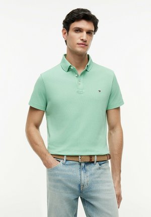 Mężczyzna o ciemnych włosach, ubrany w jasnozieloną koszulkę polo i jasnoniebieskie dżinsy z brązowym plecionym paskiem, stojący z jedną ręką w kieszeni.