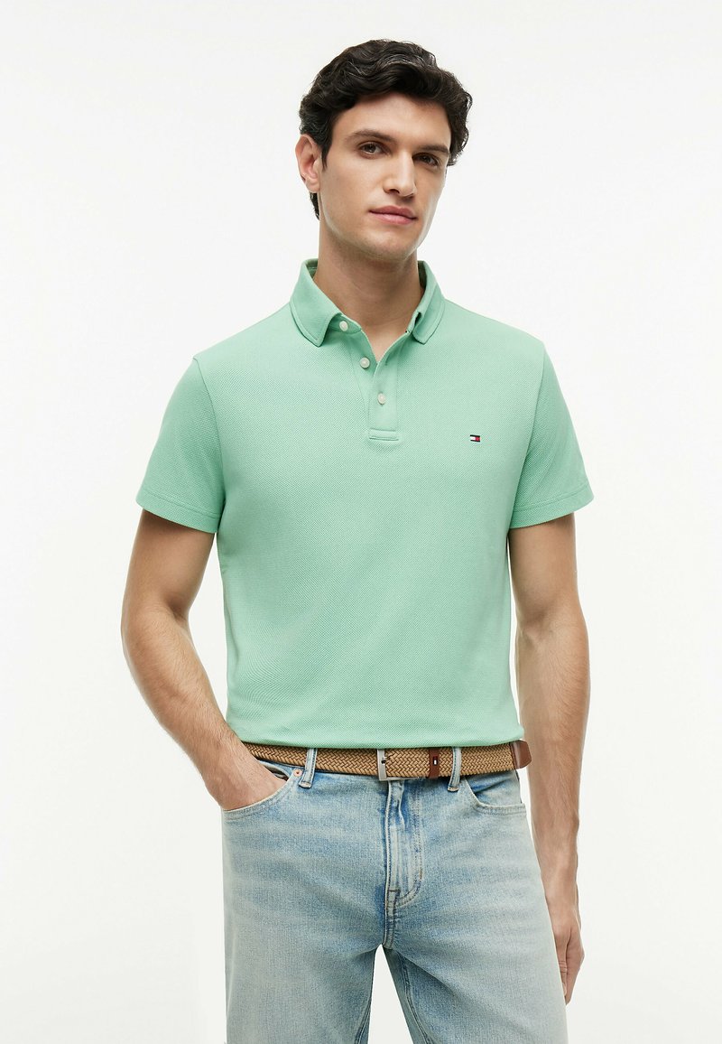 Homme aux cheveux foncés portant un polo vert clair et un jean bleu clair avec une ceinture tressée marron, debout avec une main dans la poche.