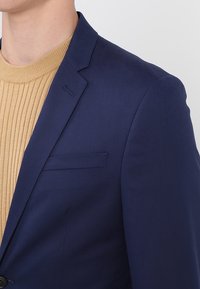 Man som bär en marinblå kavaj över en beige ribbad stickad tröja, visar detaljer om övre torso och axlar mot en enfärgad bakgrund.
