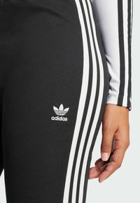 Melnas sporta legingu bikses ar baltām trim svītr�ām gar sāniem un mazu baltu Adidas logo pie gurniem, gluda auduma struktūra.
