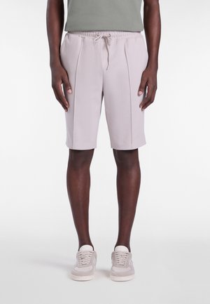Hombre vistiendo pantalones cortos hasta la rodilla de color beige claro con cintura ajustable con cordón y detalle de costura frontal, combinados con zapatillas deportivas claras.